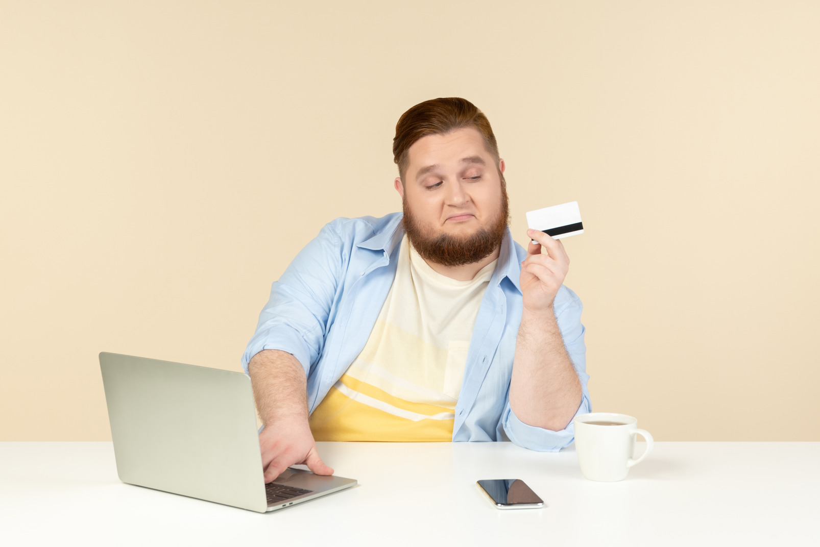 Serious looking young overweight man sitting at the home desk and holding bank card