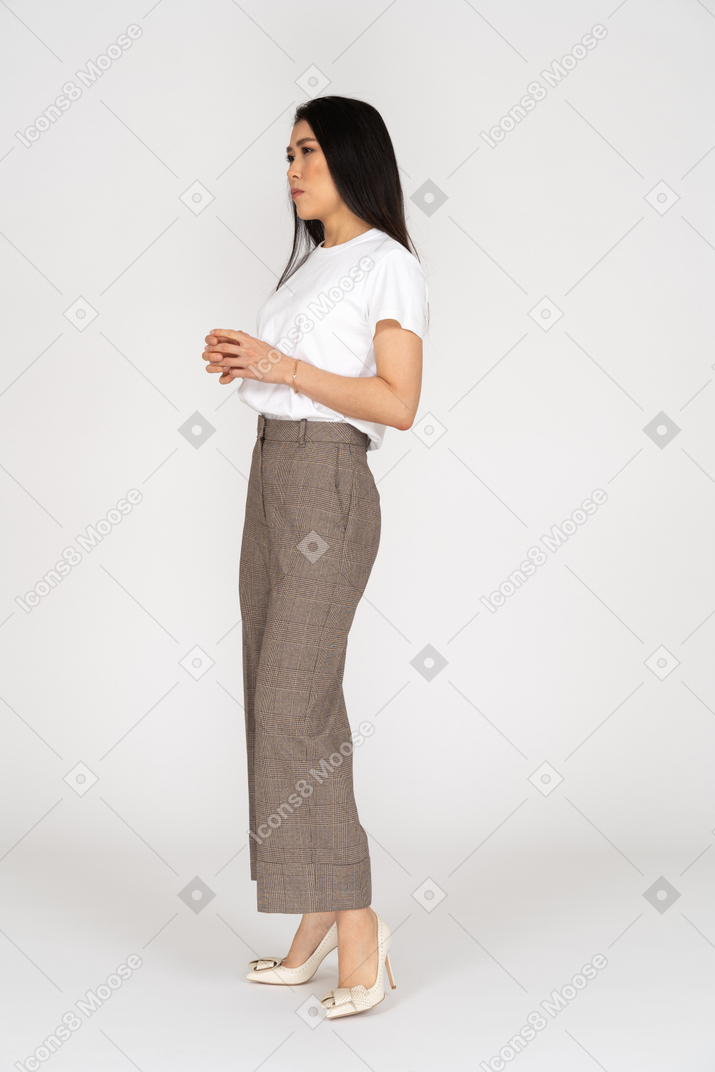Vista di tre quarti di una giovane donna seria in calzoni e maglietta che tengono le mani insieme