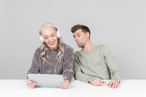 Old woman in headphones and young guy watching something on a digital tablet