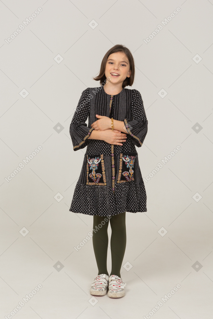一个穿着裙子的微笑小女孩的前视图，看着一边，手放在肚子上
