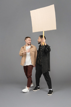 Three-quarter view of two young men carrying a billboard and chatting