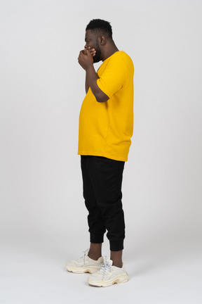 Vista laterale di un giovane uomo dalla pelle scura scioccato con una maglietta gialla che nasconde la bocca