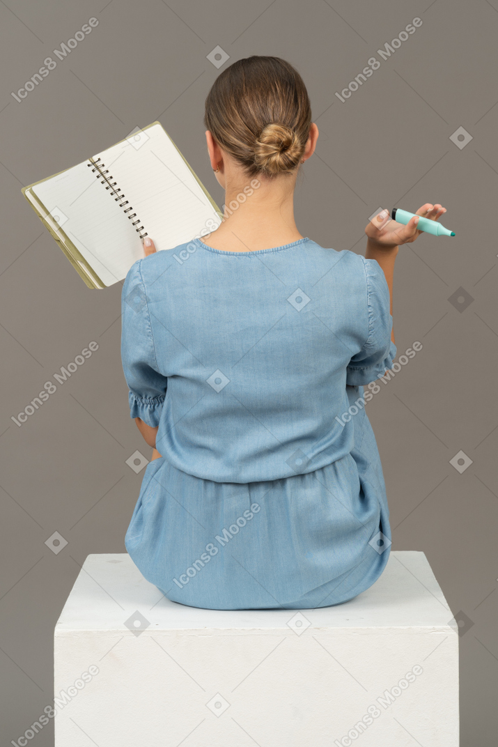 Вид сзади на молодую женщину в голубом платье, сидящую на кубе и делающую заметки