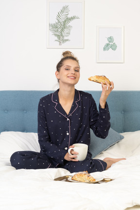 Vue de face d'une jeune femme en pyjama prenant son petit déjeuner au lit