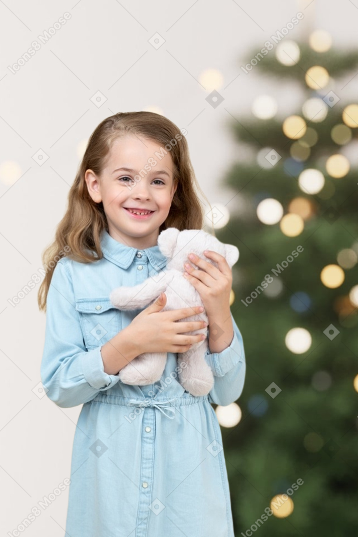 Menina segurando um ursinho de pelúcia na frente da árvore de natal