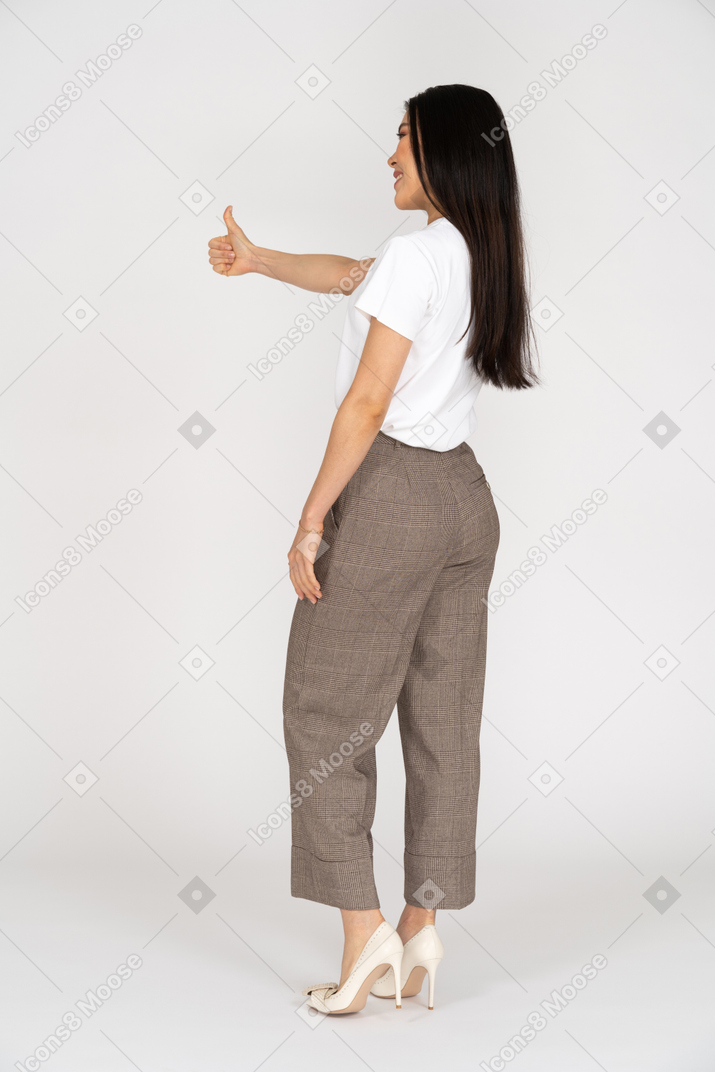 Вид сзади в три четверти улыбающейся молодой леди в бриджах и футболке, показывающей большой палец вверх