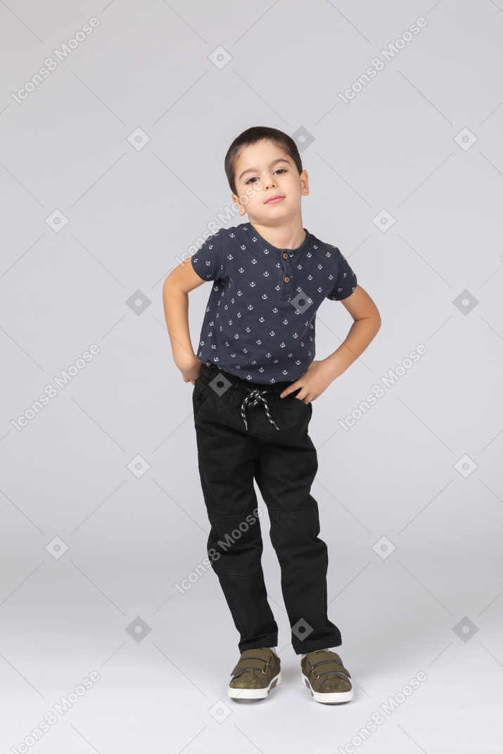 Front view of a cute boy in casual clothes posing with hands on hips and looking at camera