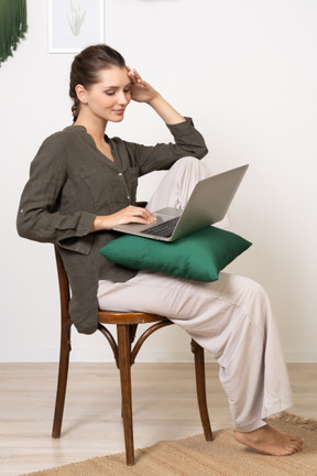 Vista de tres cuartos de una mujer joven vestida con ropa de casa sentada en una silla con una computadora portátil y tocando la frente