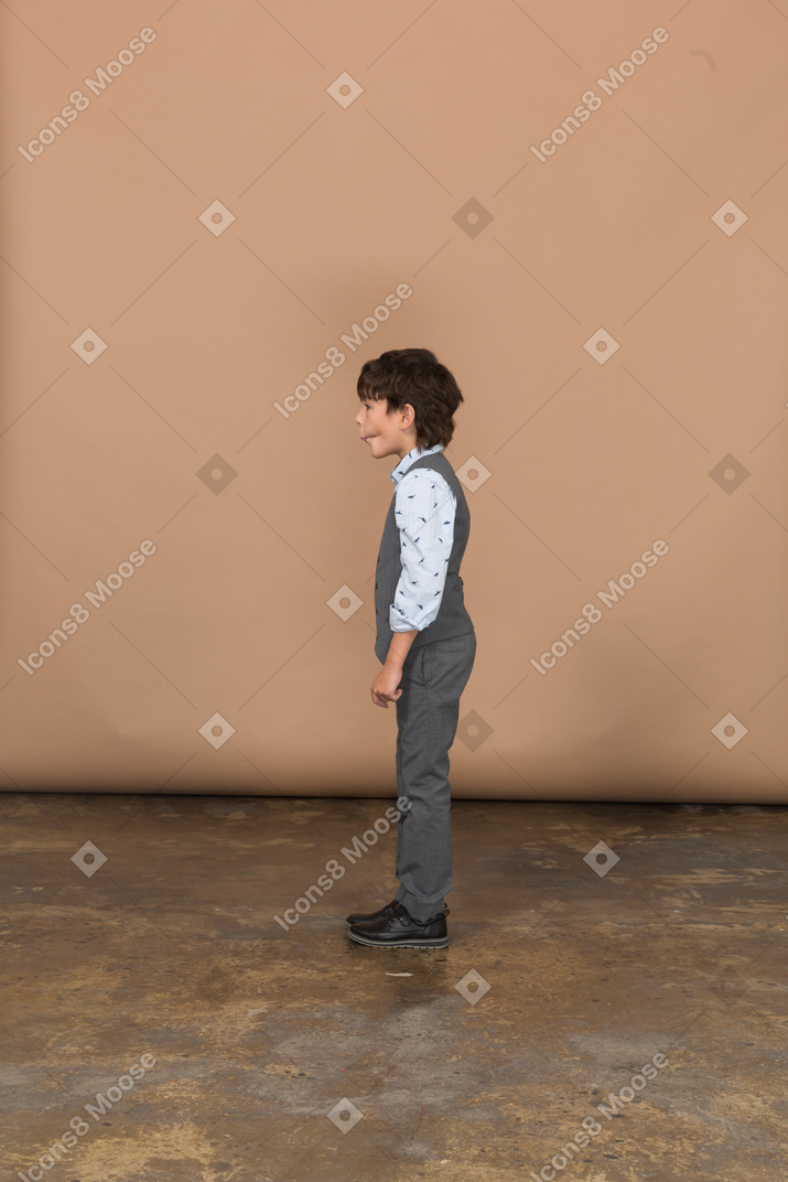 Мальчик в сером костюме стоит в профиль и корчит рожи