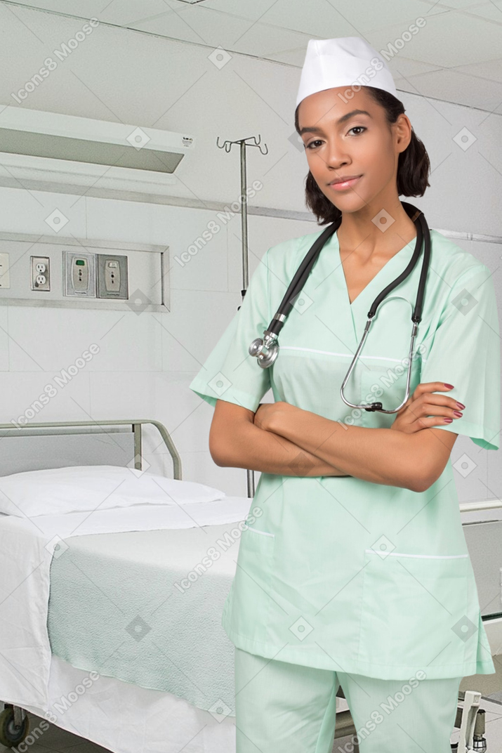 Joven doctora de pie con las manos cruzadas cerca de la cama del hospital