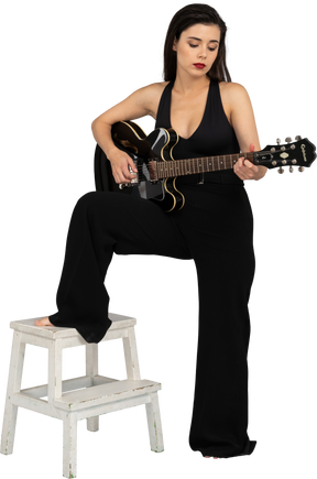 Vista frontal de uma jovem de terno preto segurando o violão e colocando a perna no banquinho