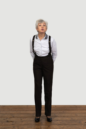 Front view of an old funny female in office clothes grimacing with her hands behind back sending a kiss