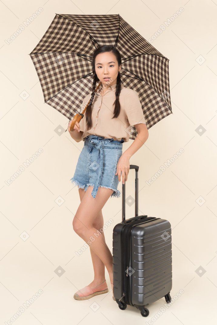 Serious young woman with umbrella and suitcase looking at camera