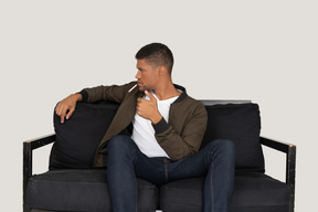Vorderansicht eines jungen arroganten mannes, der auf einem sofa sitzt und eine zigarette im mund hält