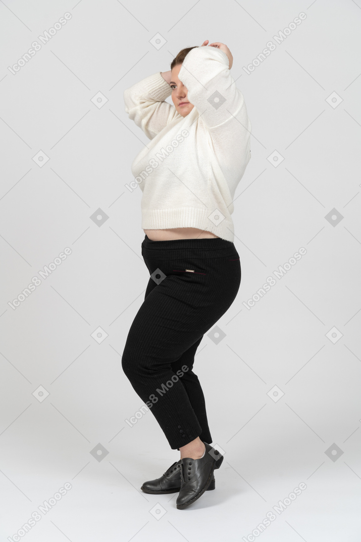Mujer regordeta en suéter blanco de pie con las manos detrás de la cabeza