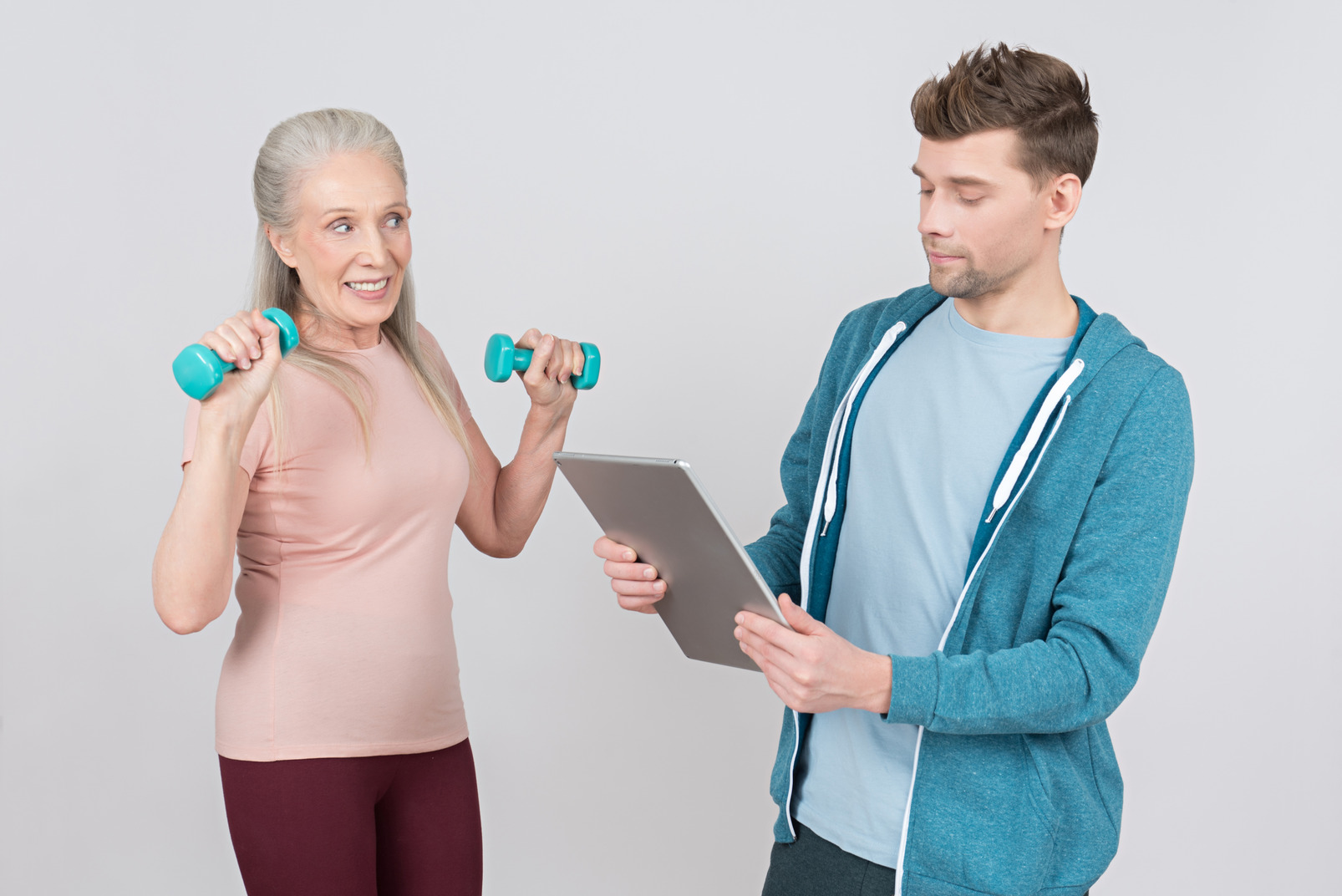 Young gym teacher looking on tablet while old woman lifting hand weights