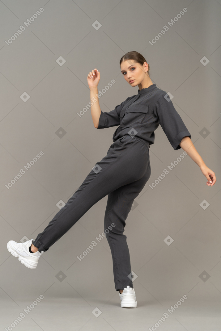 Side view of a young woman in a jumpsuit outstretching her arm & raising leg