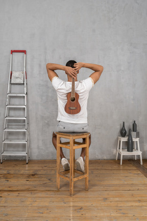 Back view of a man sitting on a stool holding an ukulele behind his back