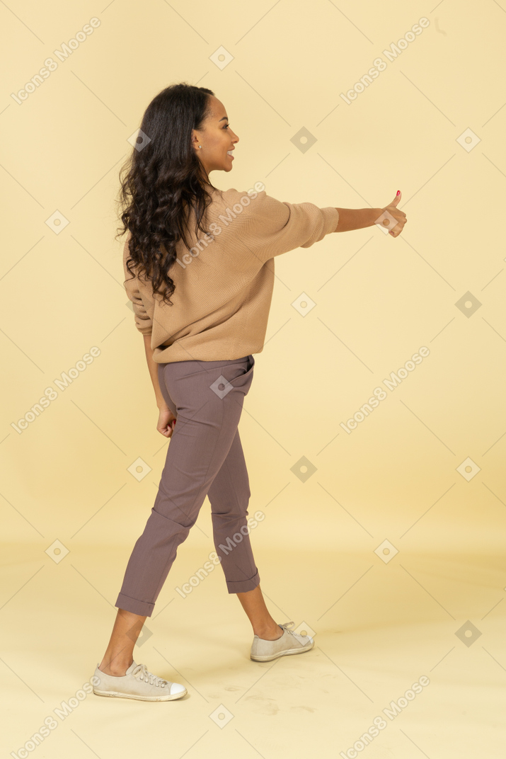 Vista posterior de tres cuartos de una mujer joven de piel oscura complacida mostrando el pulgar hacia arriba