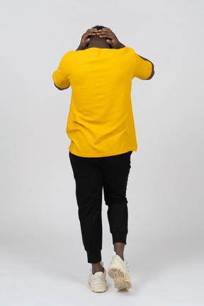 Back view of a walking young dark-skinned man in yellow t-shirt touching head