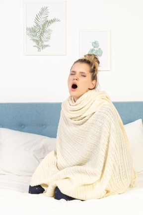 Three-quarter view of a yawning young lady wrapped in white blanket sitting in bed