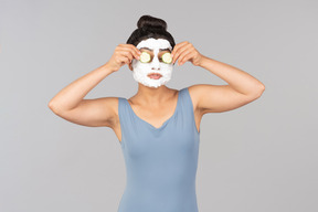Woman with white facial mask applying cucumber slices on eyes