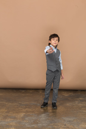 Vista frontal de um menino bonito de terno cinza em pé com o braço estendido