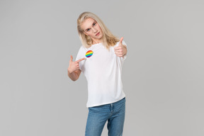 Красивый молодой человек с длинными светлыми волосами, стоящий на сером фоне, в синих джинсах и белой футболке с изображением лгбт