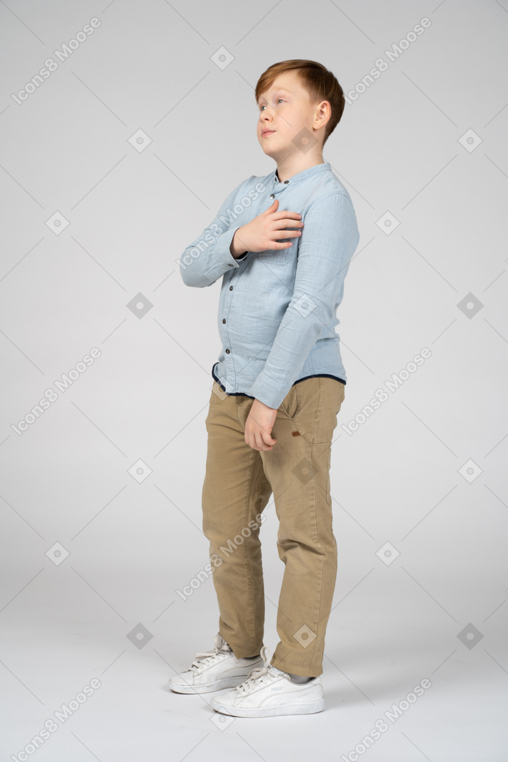 Vista lateral de um menino bonito em pé com a mão no peito e olhando para cima