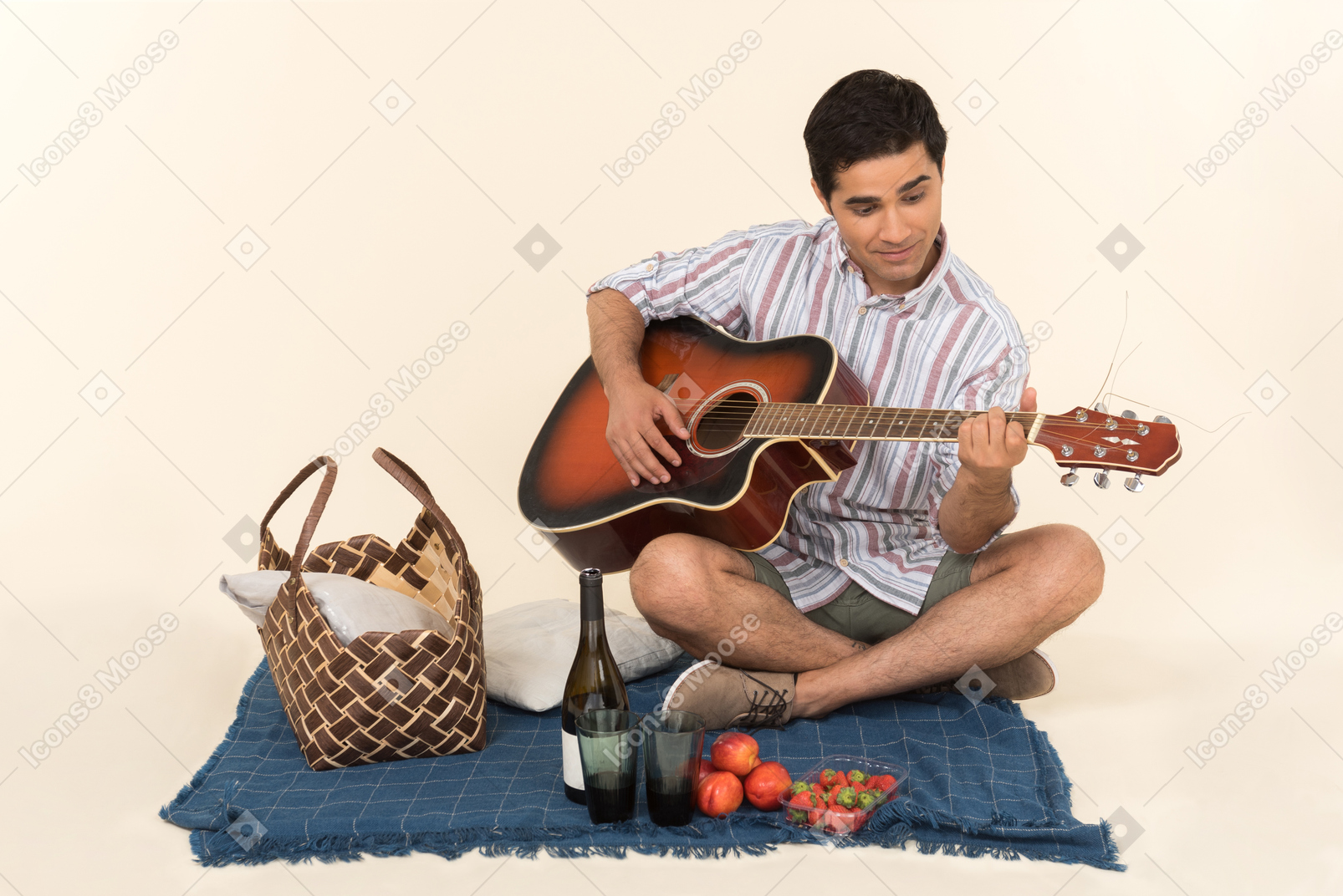 Junger kaukasischer kerl, der nahe picknickkorb auf der decke sitzt und gitarre spielt