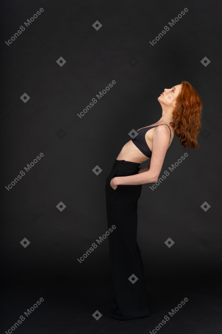 A side view of the young cute woman on the black background, holding her hands in the pockets and looking up
