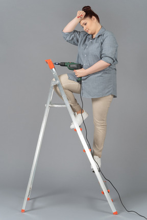 Tired young woman standing on stepladder with a drill and holding her had
