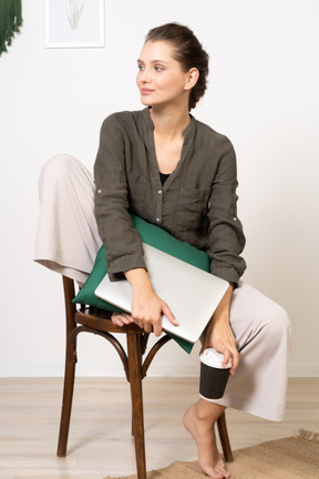Foto gratis tecnologia vista frontale di una giovane donna seduta su una sedia e con in mano il laptop e la tazza di caffè