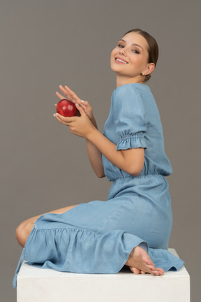 Side view of cheerful young woman with apple sitting on cube and looking at camera