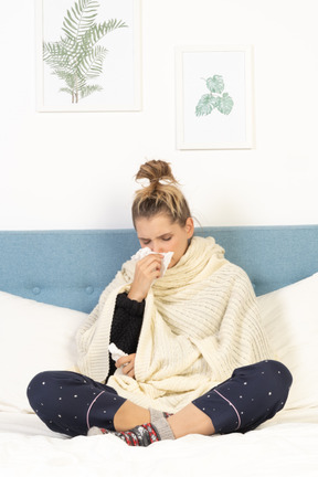 Front view of a young lady wrapped in white blanket sitting in bed and blowing nose