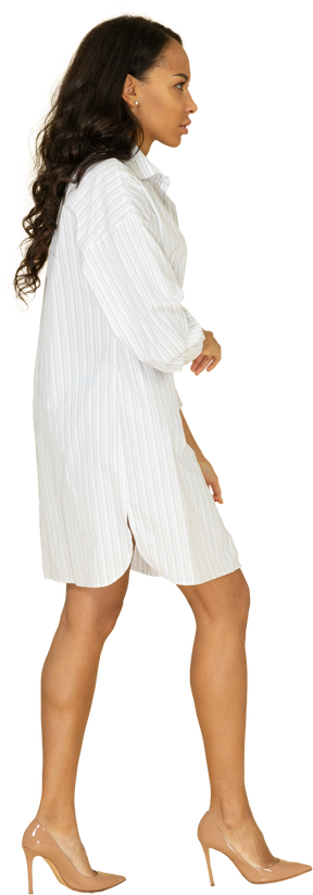 Vista lateral de una mujer joven de piel oscura caminando en vestido blanco cruzando las manos
