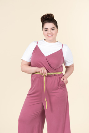 Jovem plus size em um macacão rosa, posando com uma fita métrica contra um fundo amarelo pastel