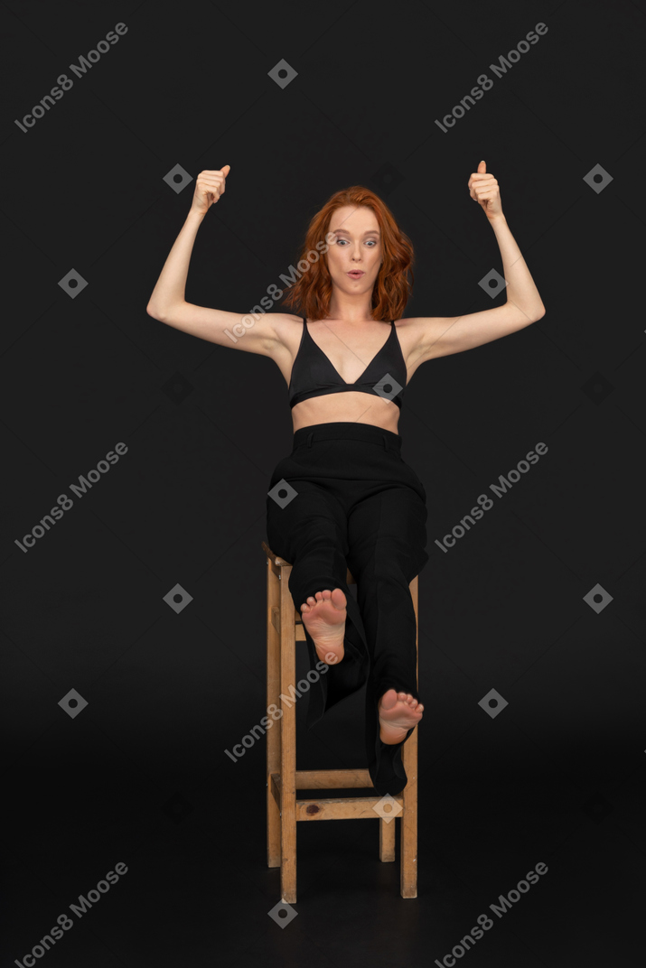 A frontal view of the young happy woman dressed in black pants and bra, sitting on the wooden chair