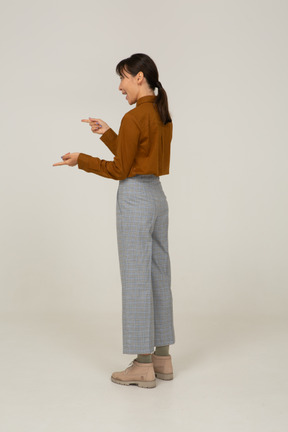 Три четверти сзади эмоциональной молодой азиатской женщины в бриджах и блузке, указывая пальцами