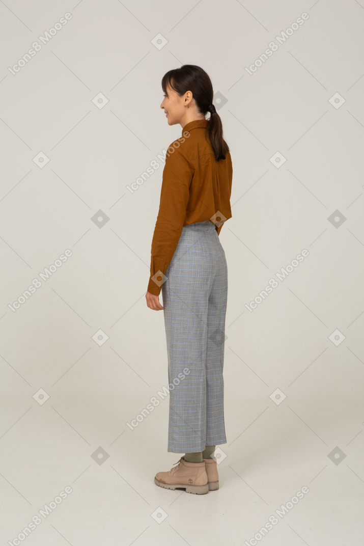 Vista traseira a três quartos de uma jovem mulher asiática sorridente, de calça e blusa, parada