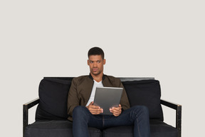 Front view of a bored young man sitting on a sofa while holding tablet