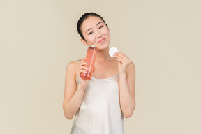 Мечтательная молодая азиатская девушка держит косметическую бутылку и ватный диск
