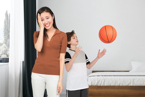 Woman holding her head and standing next to a boy playing basketball in bedroom