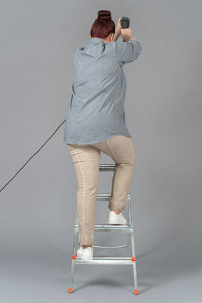 Woman standing backwards on stepladder and drilling a wall