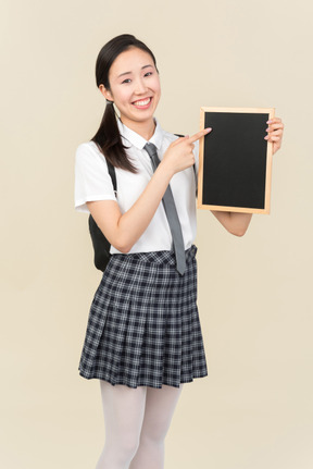 Sorrindo menina asiática da escola apontando para pequena placa preta