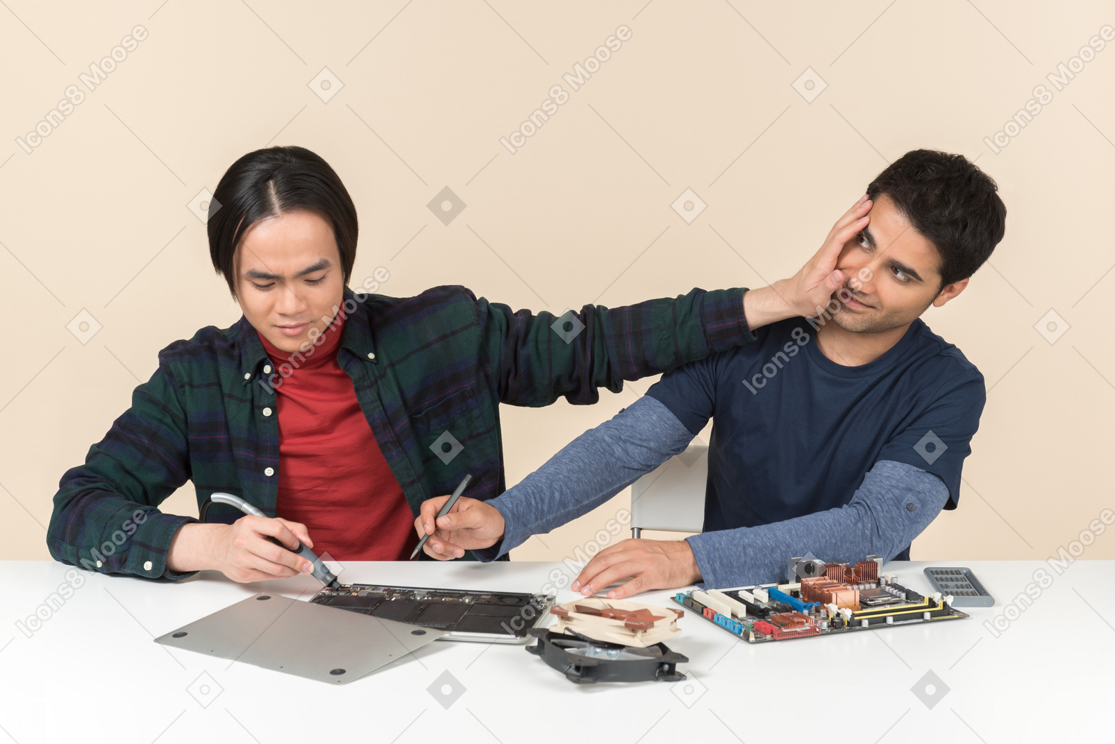 Two young geeks sitting at the table fixing something and one of them distracting other one