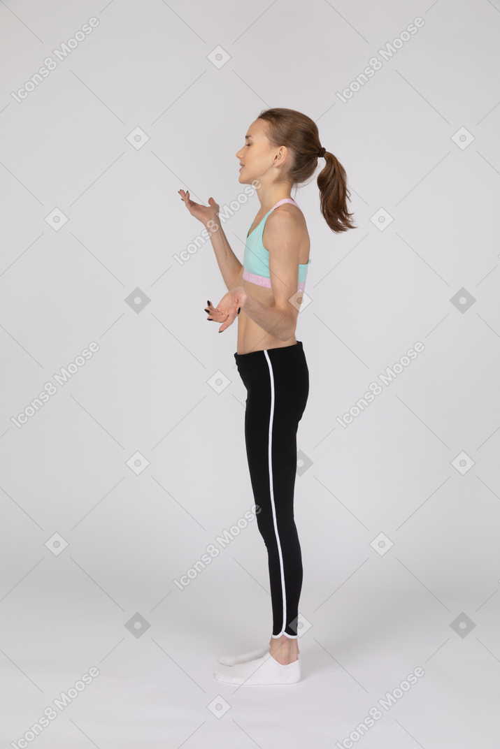 Вид сбоку на девушку-подростка в спортивной одежде, поднимающую руки, при этом что-то объясняя