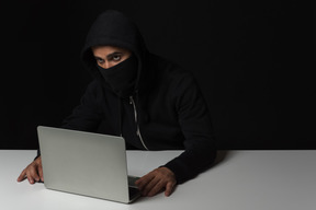 Cara de hacker sentado à mesa e trabalhando no laptop no escuro