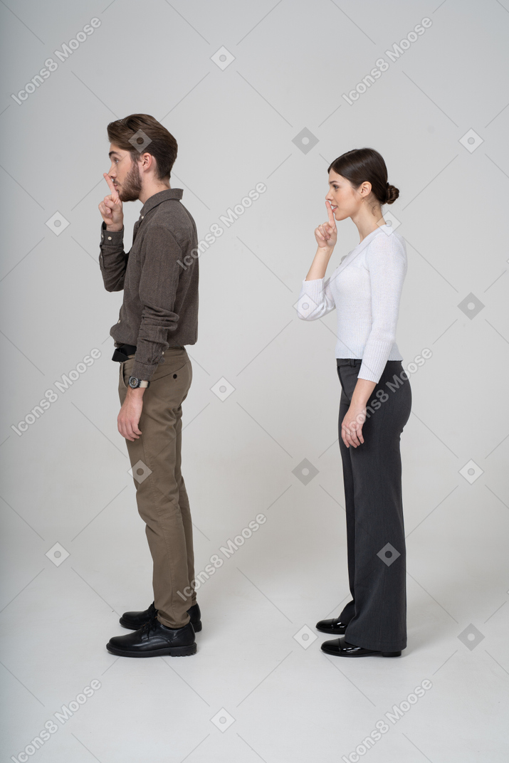 Vista lateral de um jovem casal com roupas de escritório mostrando gesto de silêncio