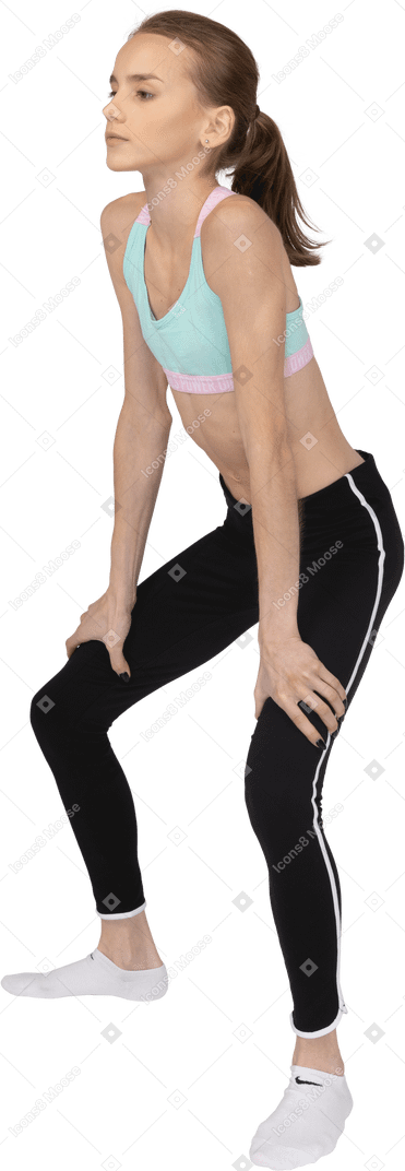 Vista de tres cuartos de una adolescente en ropa deportiva en cuclillas y poniendo las manos en las caderas